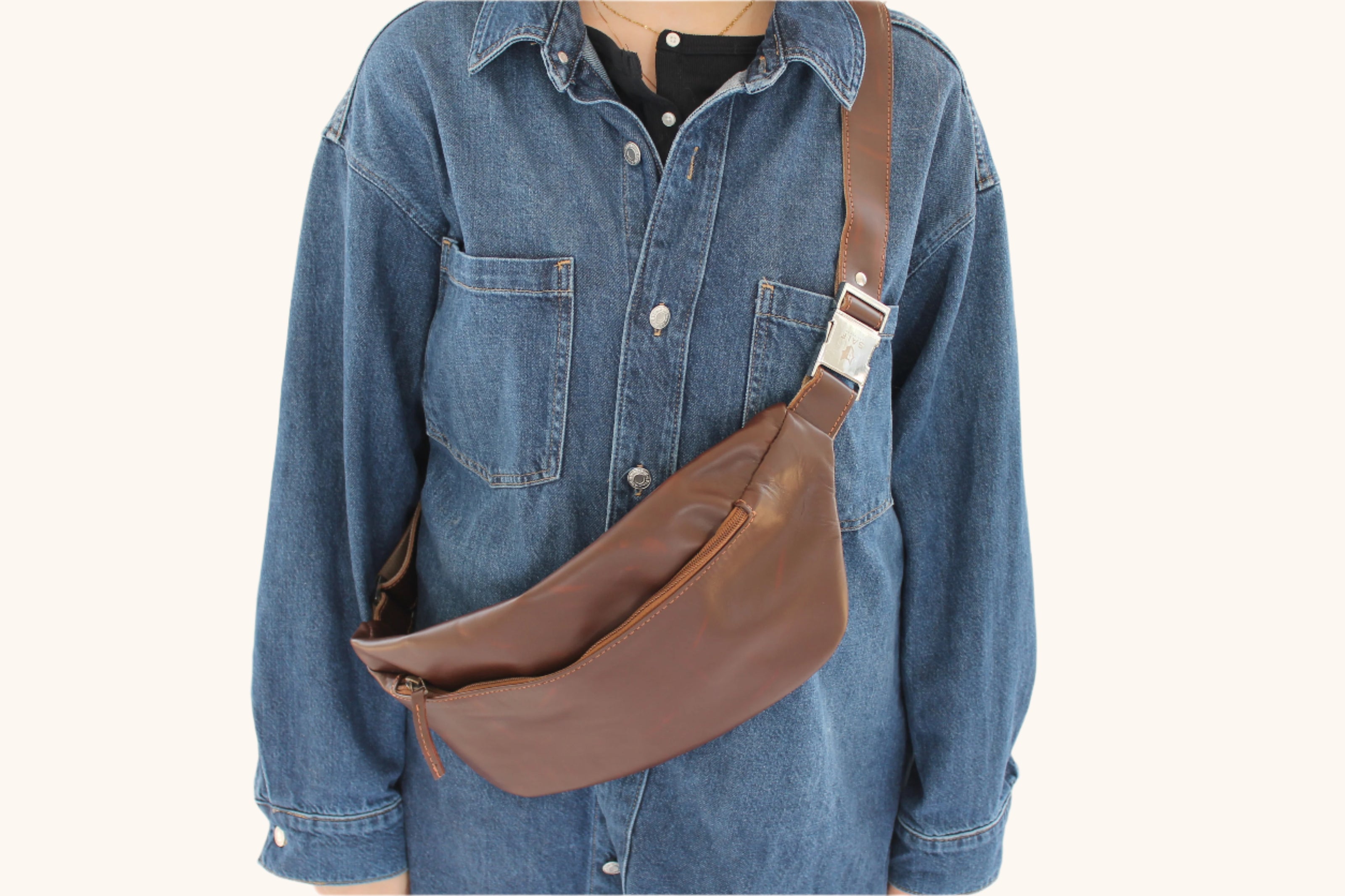 Person wearing a brown leather sling bag over a denim shirt on a white background